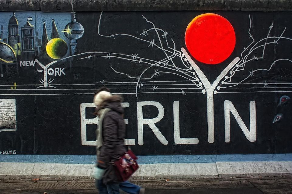 Una persona sfocata passa davanti a un grande murale di street art su un muro con la scritta 'BERLIN' in grandi lettere bianche.