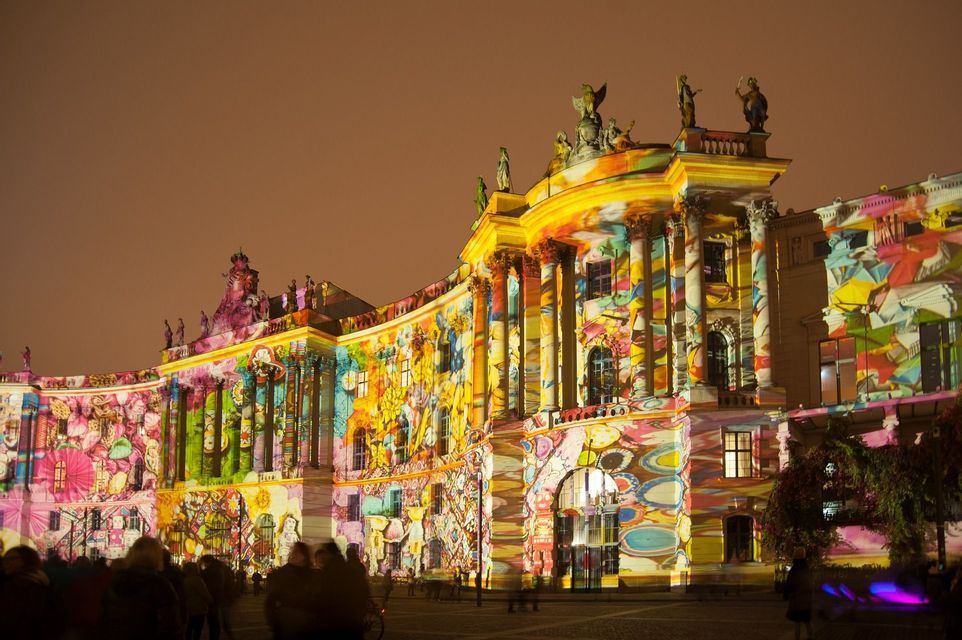 Una proiezione di luce colorata e astratta illumina di notte la facciata di un grande edificio classico, con spettatori in primo piano.