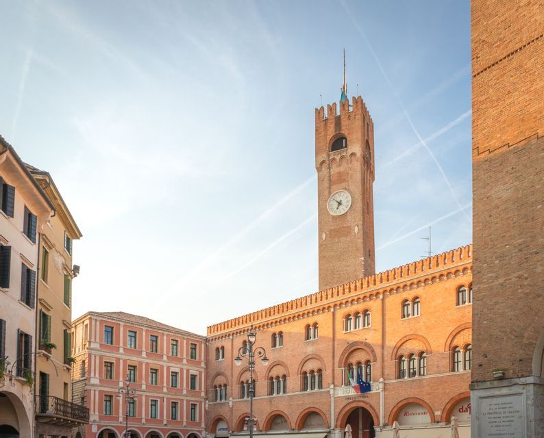 A sunlit, red brick clock tower and an adjacent arched building stand in a historic town square under a pale blue sky.