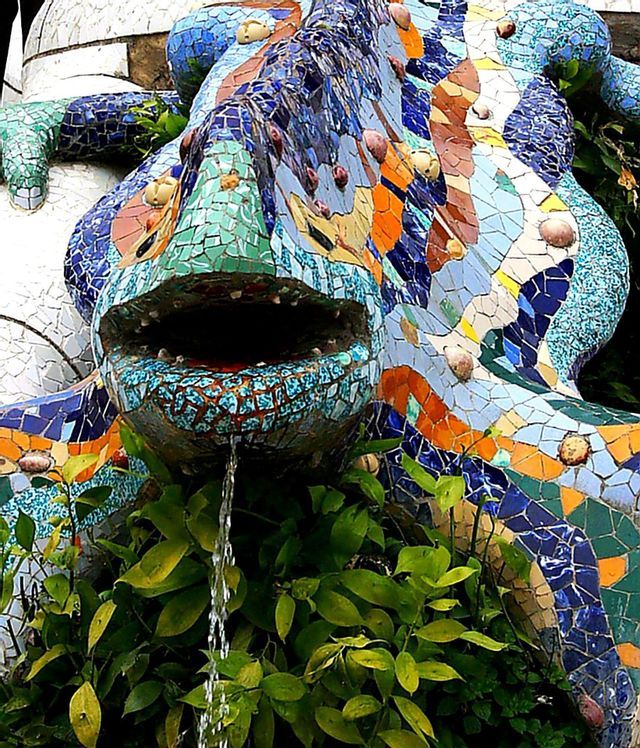 Primo piano di una scultura a forma di lucertola a mosaico colorata che funge da fontana, con acqua che sgorga dalla sua bocca aperta su piante verdi.