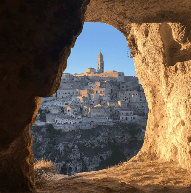 Un antico borgo in pietra adagiato su un fianco di collina, con un campanile, visto attraverso l'apertura di una grotta durante l'ora d'oro.