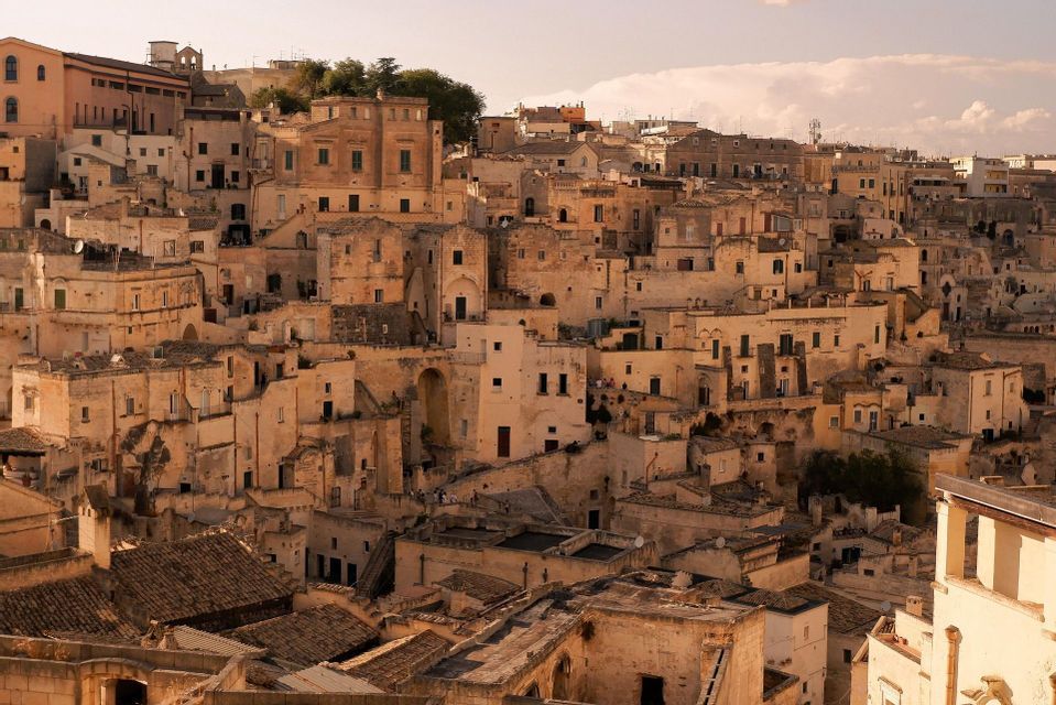 Un fitto paesaggio urbano di antichi edifici in pietra color sabbia, costruiti fittamente su una collina sotto un cielo velato.
