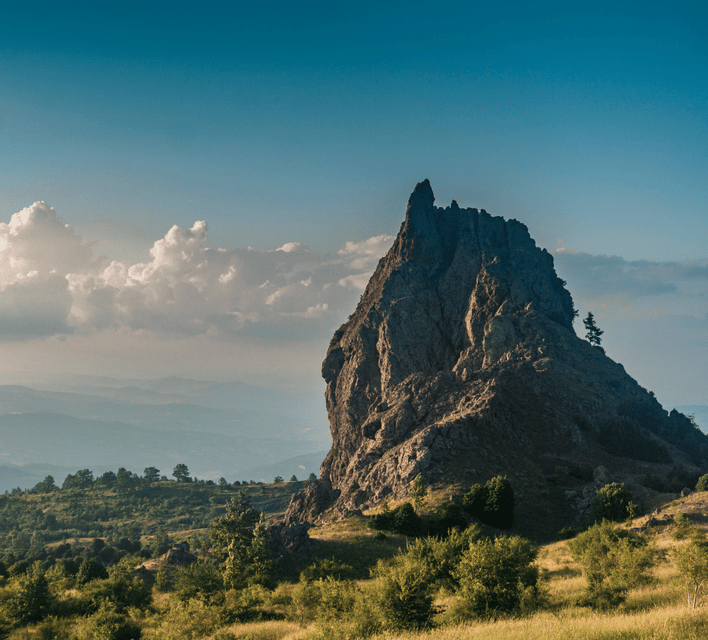 Una formazione rocciosa frastagliata si erge imponente su un paesaggio verde, con montagne sfumate all'orizzonte sotto un cielo parzialmente nuvoloso.