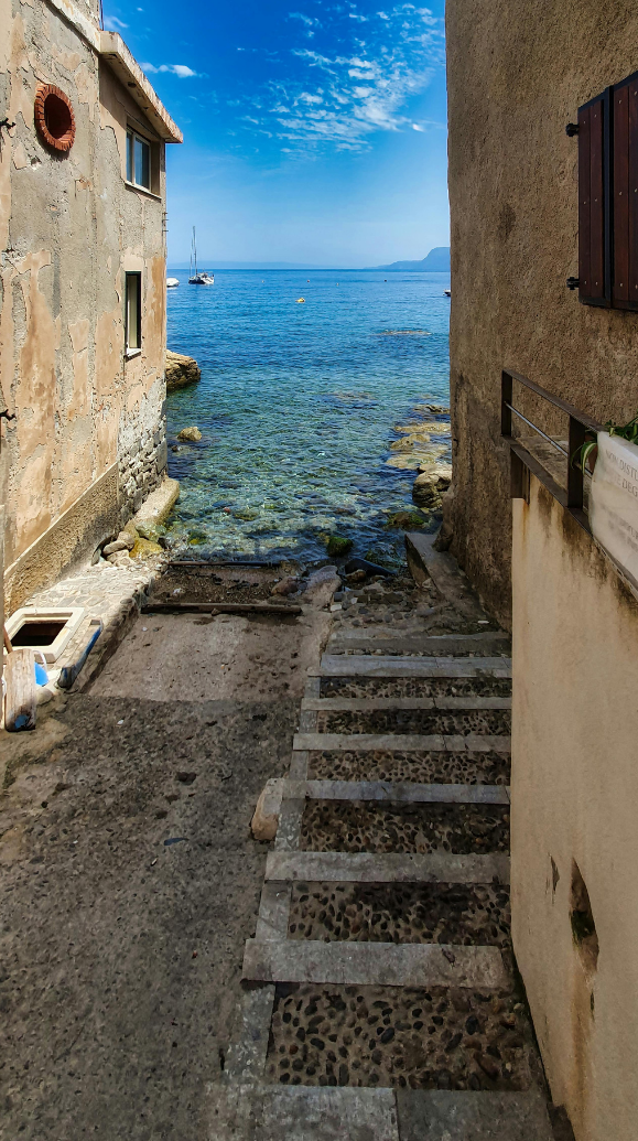 Una scalinata in pietra scende attraverso un vicolo stretto tra due edifici, conducendo al mare azzurro e limpido.