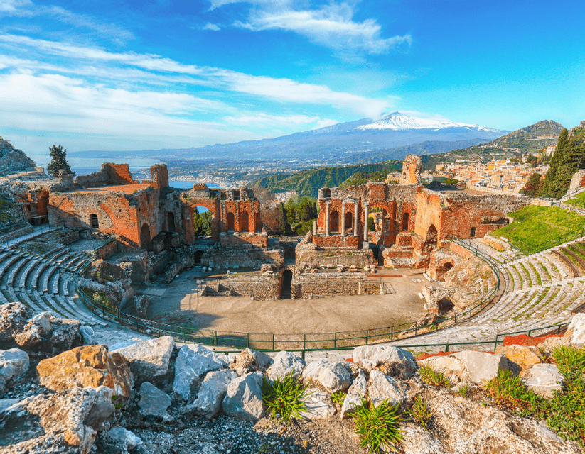 Un'ampia veduta delle rovine di un antico anfiteatro, con una città costiera e un vulcano innevato sullo sfondo.
