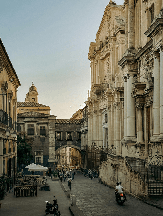 Una strada stretta in una città storica con palazzi ornati, un caffè all'aperto e gente che passeggia.