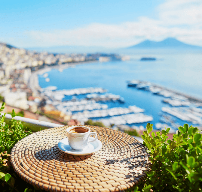Una tazza di espresso su un tavolino di vimini che si affaccia su una baia costiera assolata con un porticciolo e una montagna distante.