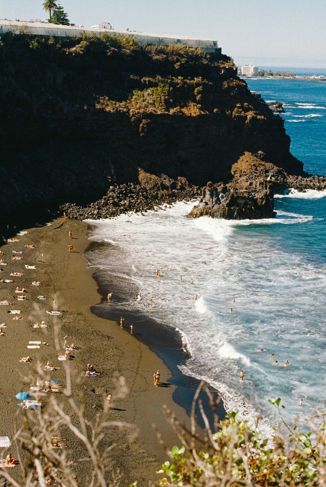 Una vista dall'alto di una spiaggia di sabbia nera dove le persone nuotano nell'oceano e prendono il sole accanto a una grande scogliera rocciosa.