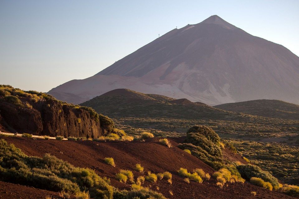 Un grande vulcano si erge su un paesaggio di terra vulcanica rossa e arbusti verdi sotto un cielo pallido e limpido.