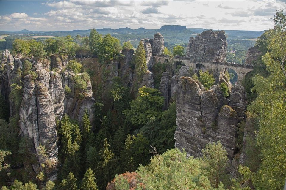 Eine lange Steinbrücke mit Bögen überquert ein von hohen, baumbewachsenen Felsformationen umgebenes Gebirgstal.