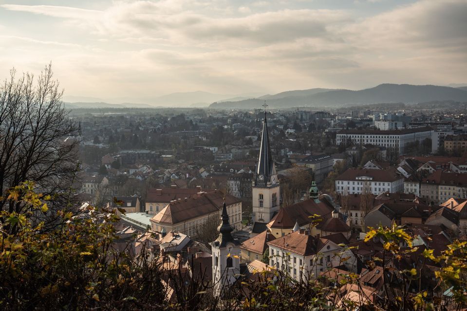 Eine Stadtansicht mit einem markanten Kirchturm und rotgedeckten Gebäuden unter einem bewölkten Himmel, mit Bergen in der Ferne.