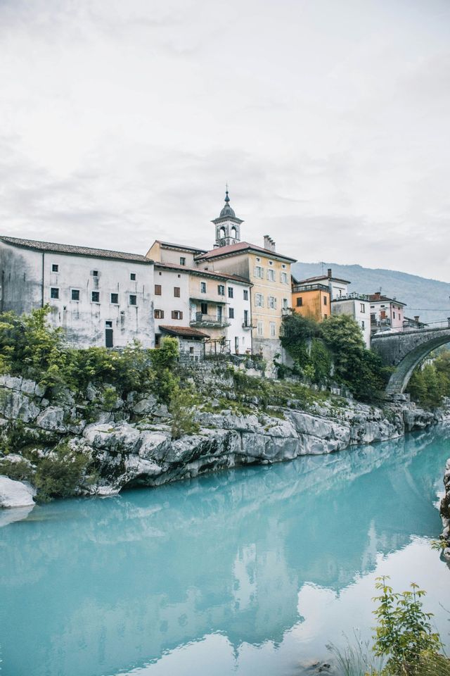 Eine Stadt mit Glockenturm liegt auf einer felsigen Klippe über einem helltürkisen Fluss und einer Steinbrücke.