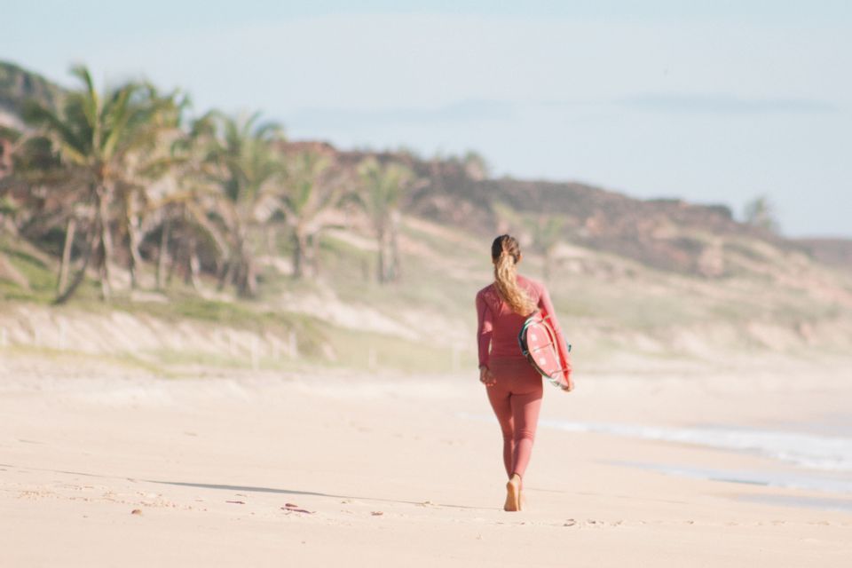 Una mujer en traje de neopreno rosa, vista de espaldas, camina por una playa de arena llevando una tabla de surf.