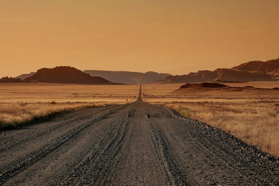 Une longue route de gravier traverse un paysage désertique vers des montagnes lointaines sous un ciel chaud et orangé.