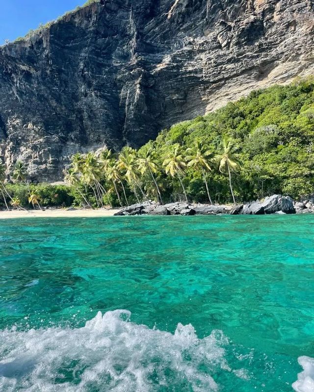 Una vista dalla scia di una barca attraverso acque turchesi verso una spiaggia sabbiosa fiancheggiata da palme alla base di una grande scogliera rocciosa.