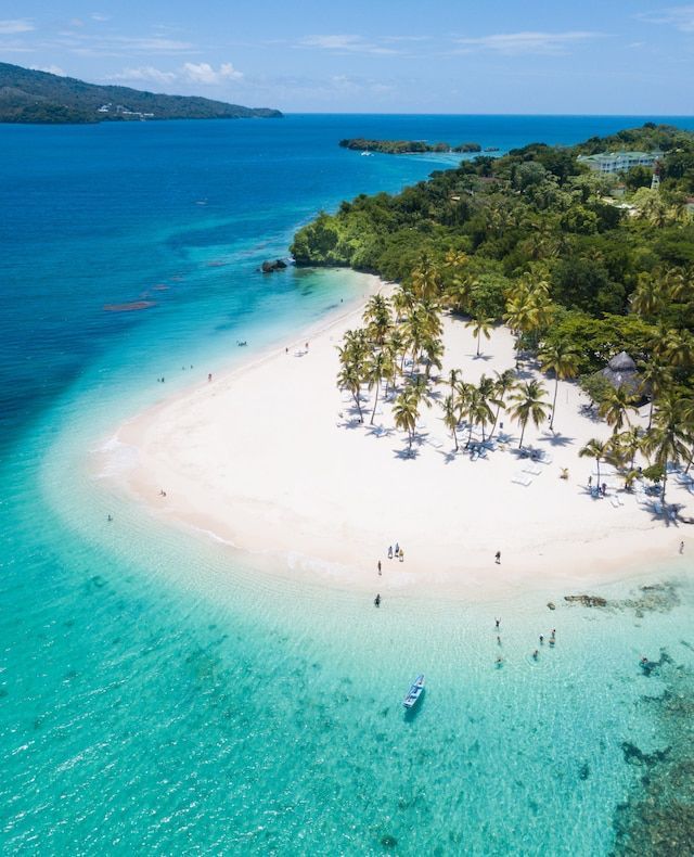 Una veduta aerea di una spiaggia tropicale di sabbia bianca con palme, circondata da acque turchesi cristalline con persone che nuotano.
