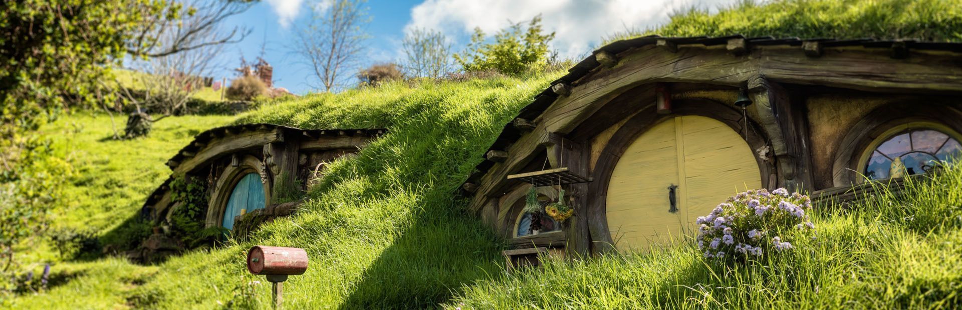 Deux maisons fantaisistes avec des portes rondes sont intégrées dans une colline verdoyante sous un ciel ensoleillé.