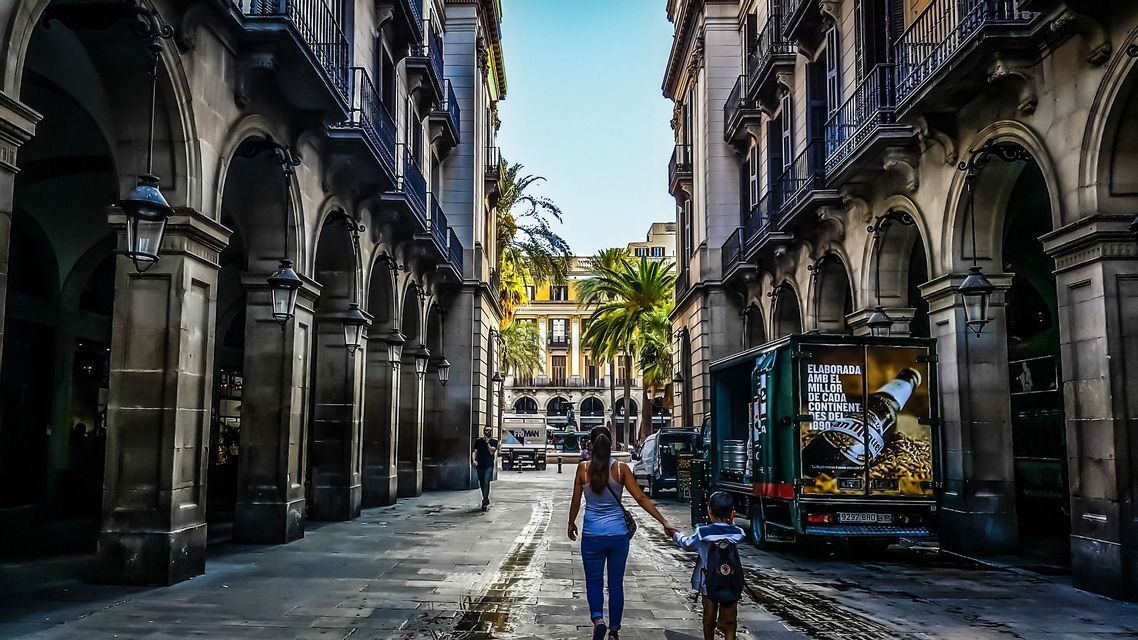 Una donna e un bambino si tengono per mano camminando lungo una strada stretta fiancheggiata da colonnati ad arco verso una piazza illuminata dal sole.
