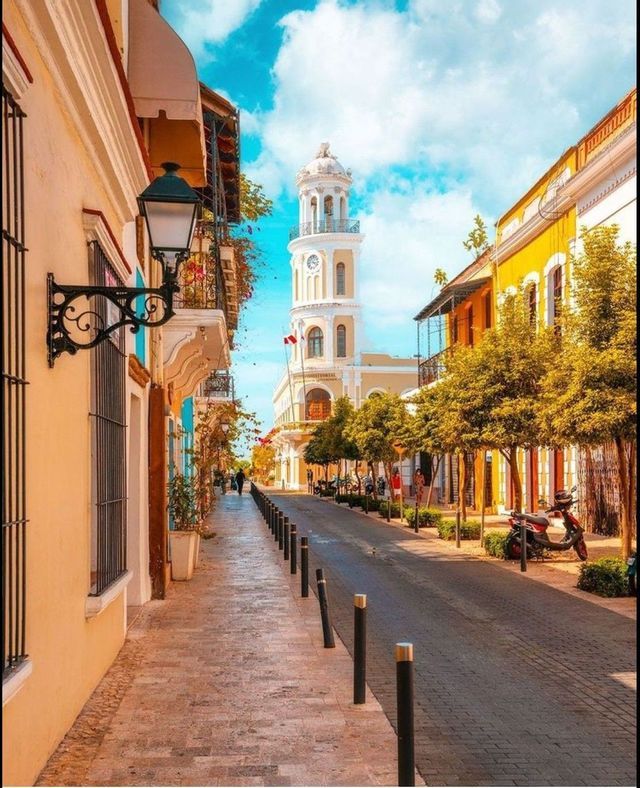 Una stretta strada acciottolata fiancheggiata da edifici colorati conduce a una torre dell'orologio bianca sotto un cielo blu con nuvole.