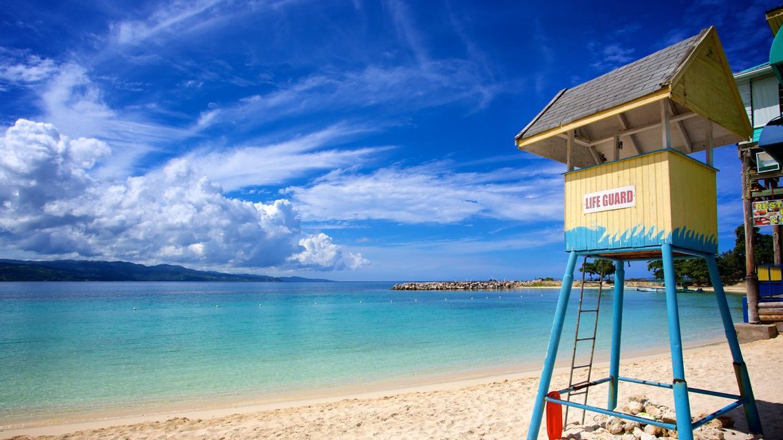 Una torre de salvavidas amarilla y azul se alza en una playa de arena junto a aguas tranquilas de color turquesa bajo un cielo parcialmente nublado.