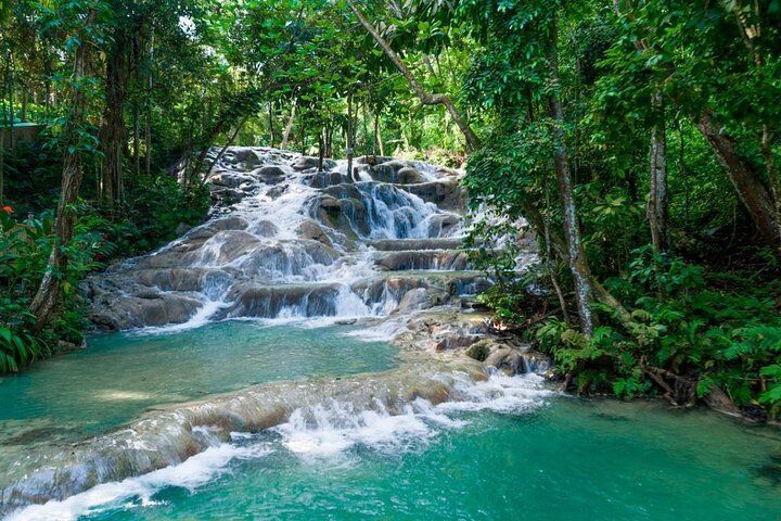 Una cascada escalonada fluye sobre rocas pulidas hacia una poza turquesa, rodeada por un frondoso bosque tropical.