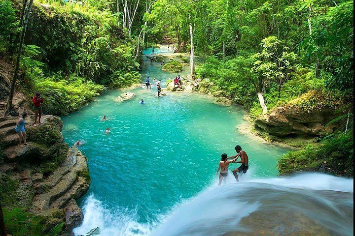 Un grupo de WeRoad disfruta de una cascada selvática de color turquesa, con algunos bañándose abajo y dos de pie al borde del salto de agua.