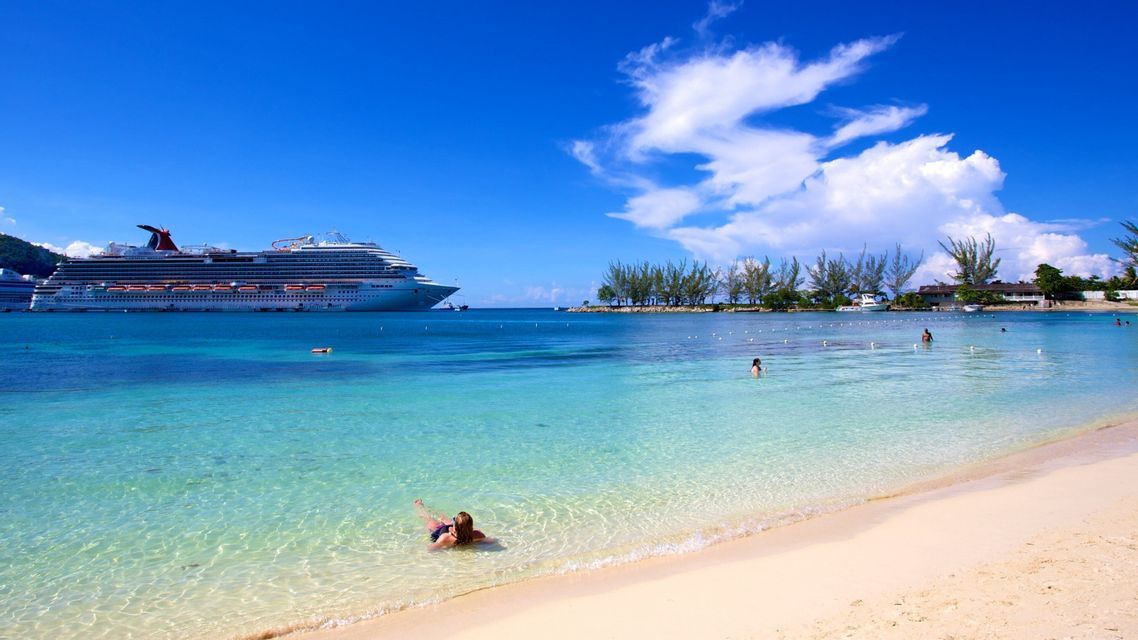 Un crucero anclado en aguas turquesas cerca de una playa de arena con gente nadando bajo un cielo azul y soleado.