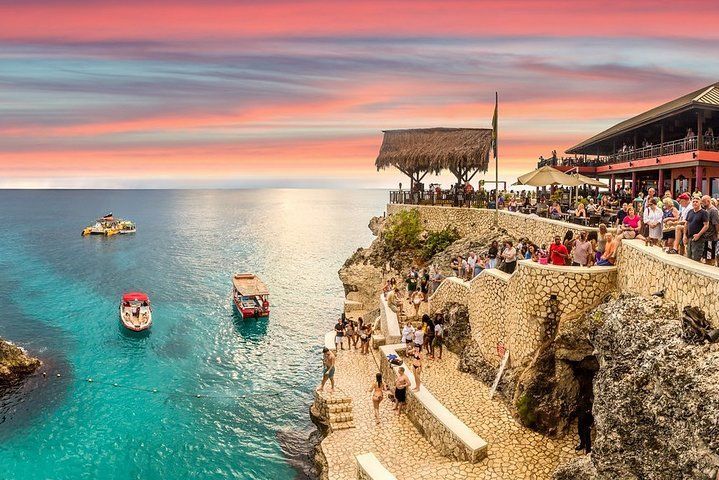 La gente se reúne en un acantilado de piedra con un restaurante, observando barcos en el agua turquesa durante una colorida puesta de sol.