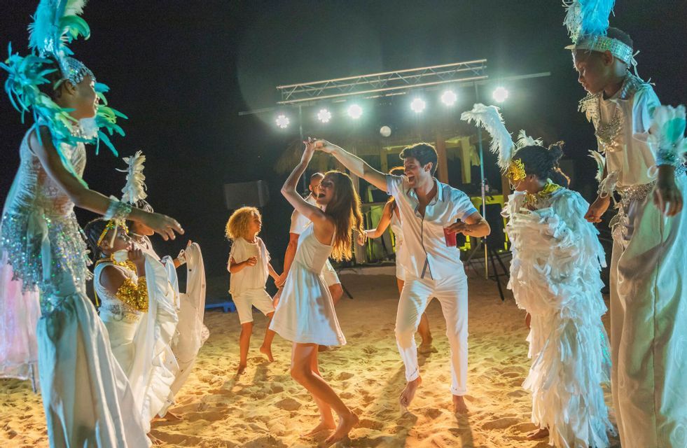 Un viaje en grupo de WeRoad bailando en la arena por la noche durante una fiesta en la playa con artistas en elaborados disfraces de plumas.
