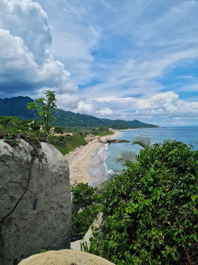 Una veduta dall'alto da uno sperone roccioso mostra una spiaggia sabbiosa che si estende lungo una costa boscosa sotto un cielo parzialmente nuvoloso.