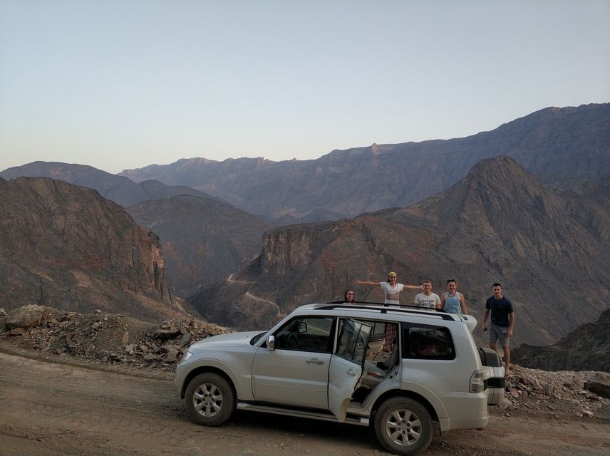 Un viaggio di gruppo WeRoad posa accanto a un SUV bianco su una strada sterrata che si affaccia su una vasta catena montuosa rocciosa