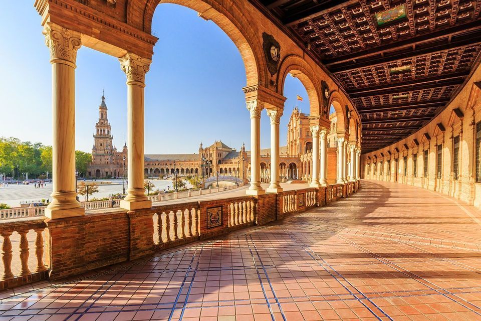 Vue d'une arcade ensoleillée avec colonnes et arches, surplombant une grande place et un bâtiment historique.