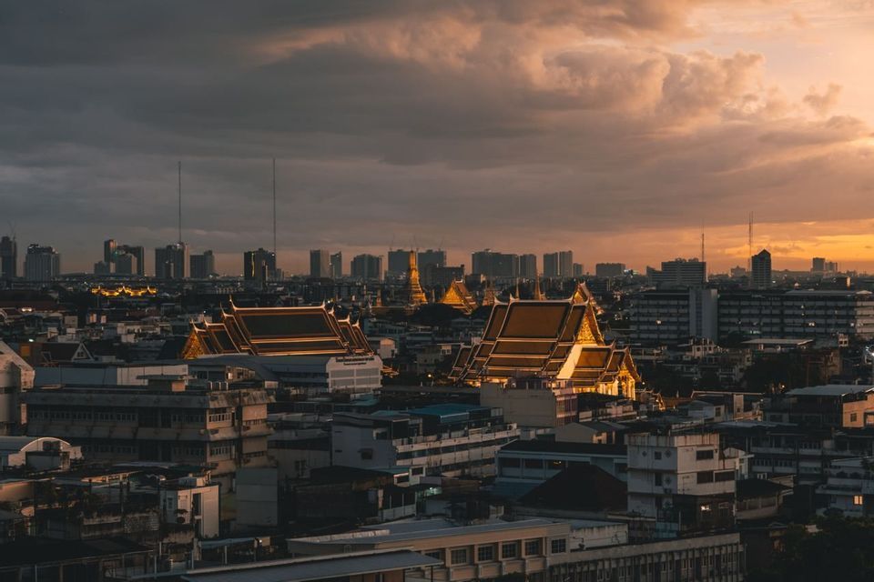Illuminated temple roofs stand out against a dense cityscape with a modern skyline at sunset under a cloudy sky.
