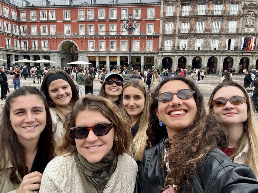 Un voyage de groupe WeRoad de femmes souriantes prenant un selfie sur une place animée d'une ville européenne avec des bâtiments historiques.