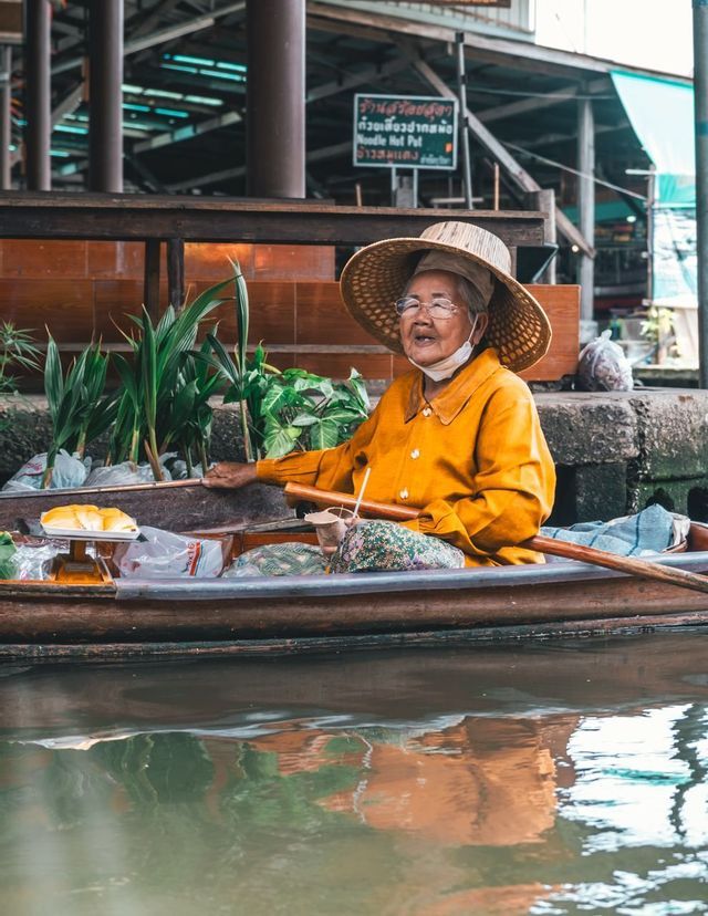 Una venditrice anziana, con una camicia gialla e un grande cappello di paglia, siede in una barca di legno colma di merci sull'acqua.