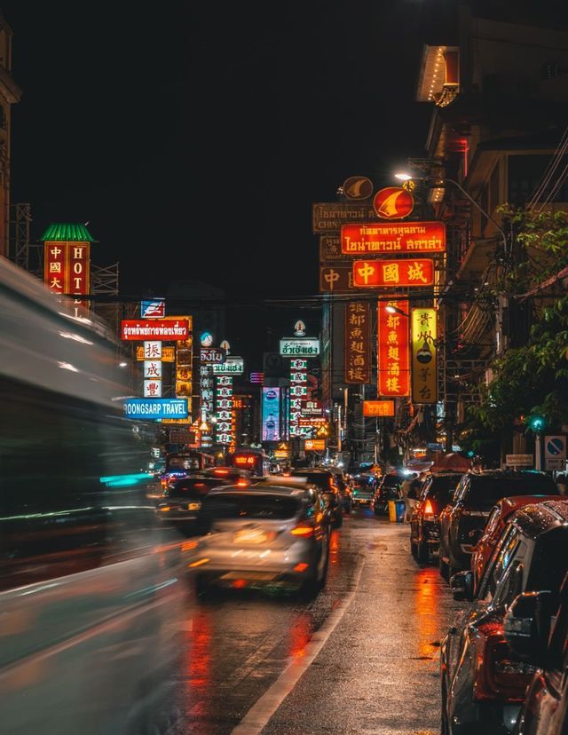 Eine belebte Stadtstraße bei Nacht, erfüllt von Autoverkehr und beleuchtet von farbigen Neonschildern, die sich auf dem nassen Pflaster spiegeln.