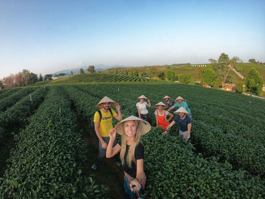 Eine WeRoad-Gruppe mit konischen Hüten lächelt für ein Selfie zwischen den sanften grünen Hügeln einer Teeplantage.