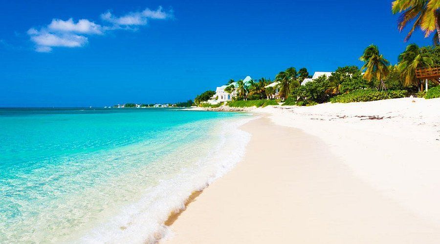 Ein weißer Sandstrand mit klarem, türkisfarbenem Wasser, das ans Ufer plätschert, gesäumt von Palmen und Villen unter einem strahlend blauen Himmel.
