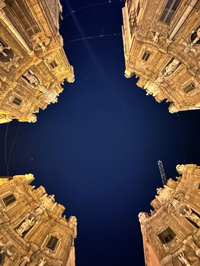 Una prospettiva dal basso di quattro angoli di palazzi sfarzosi illuminati che incorniciano un cielo notturno buio e stellato.