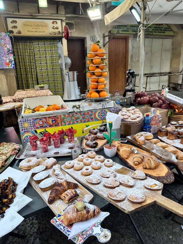 Una bancarella del mercato espone dolci freschi come ciambelle e cornetti, accanto a una pila di arance e uno spremiagrumi.