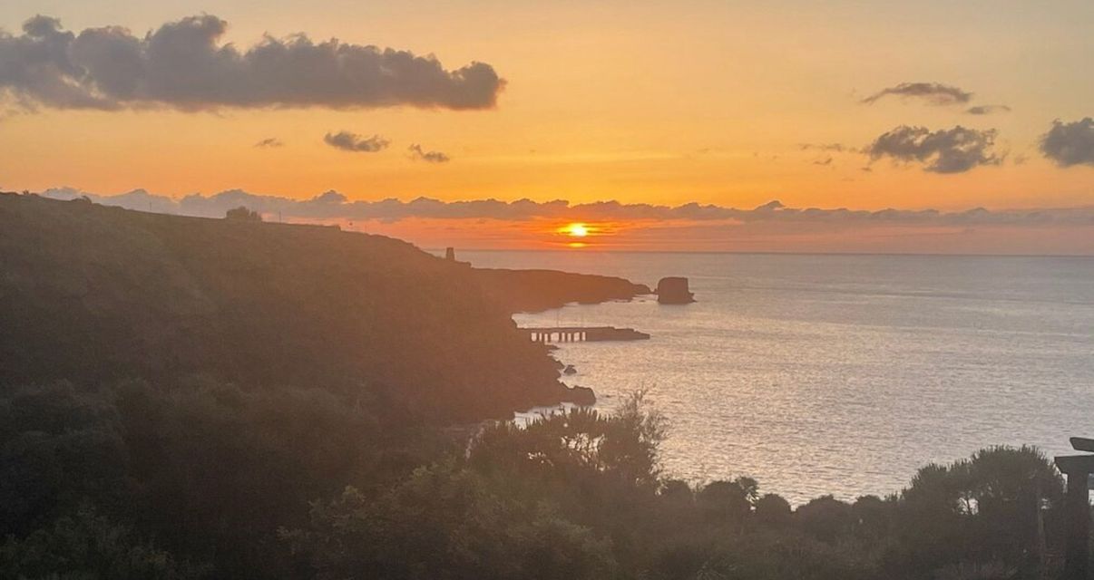 Un tramonto arancione sull'oceano accanto a una costa collinare con un piccolo molo e nuvole sparse.