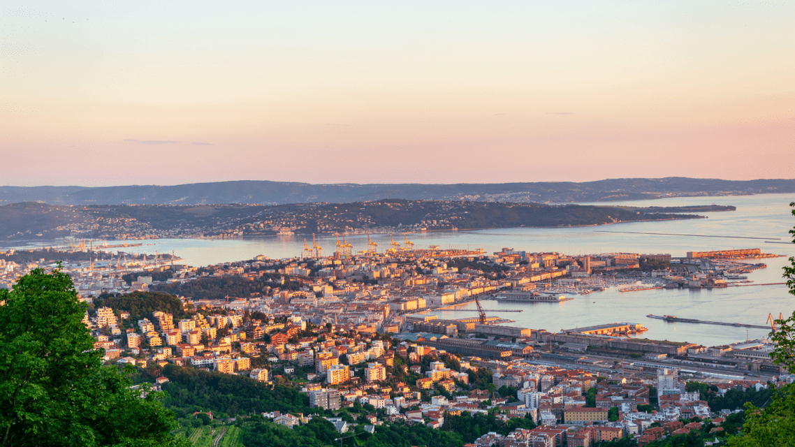 Una vista dall'alto di una città costiera e del suo porto, illuminata dalla luce calda del tramonto, con colline che circondano la baia.