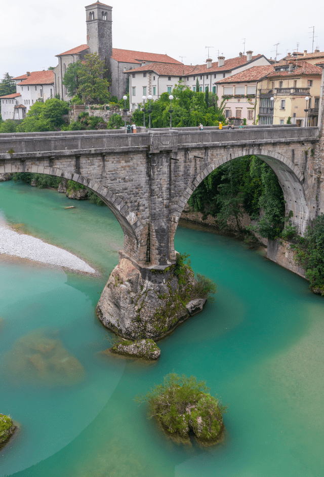 Un ponte storico in pietra si staglia su un fiume turchese, con un centro storico e un campanile visibili sulla riva opposta sotto un cielo nuvoloso.