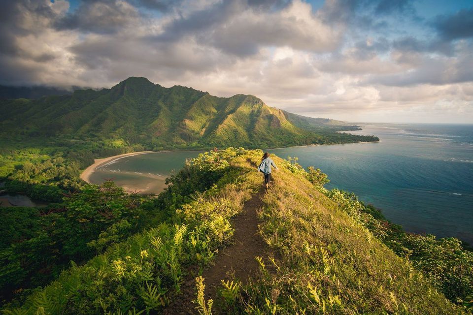 Eine Person geht auf einem Feldweg entlang eines grünen Bergrückens mit Blick auf das Meer, eine sandige Bucht und Berge unter bewölktem Himmel.