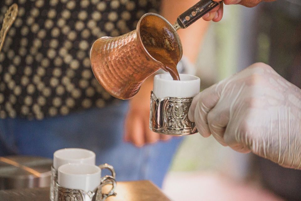 Una persona vierte café tradicional de una cafetera de cobre en una pequeña taza ornamentada sostenida por una mano enguantada.
