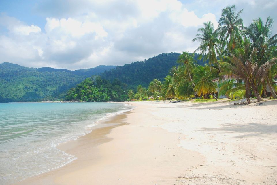 Una spiaggia di sabbia bianca con acqua turchese, fiancheggiata da palme di fronte a verdi colline lussureggianti sotto un cielo parzialmente nuvoloso.