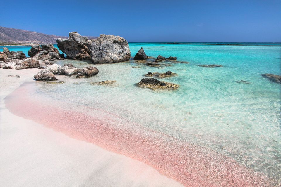 Sabbia rosa su una spiaggia bianca incontra acque turchesi cristalline con grandi rocce sotto un cielo blu.