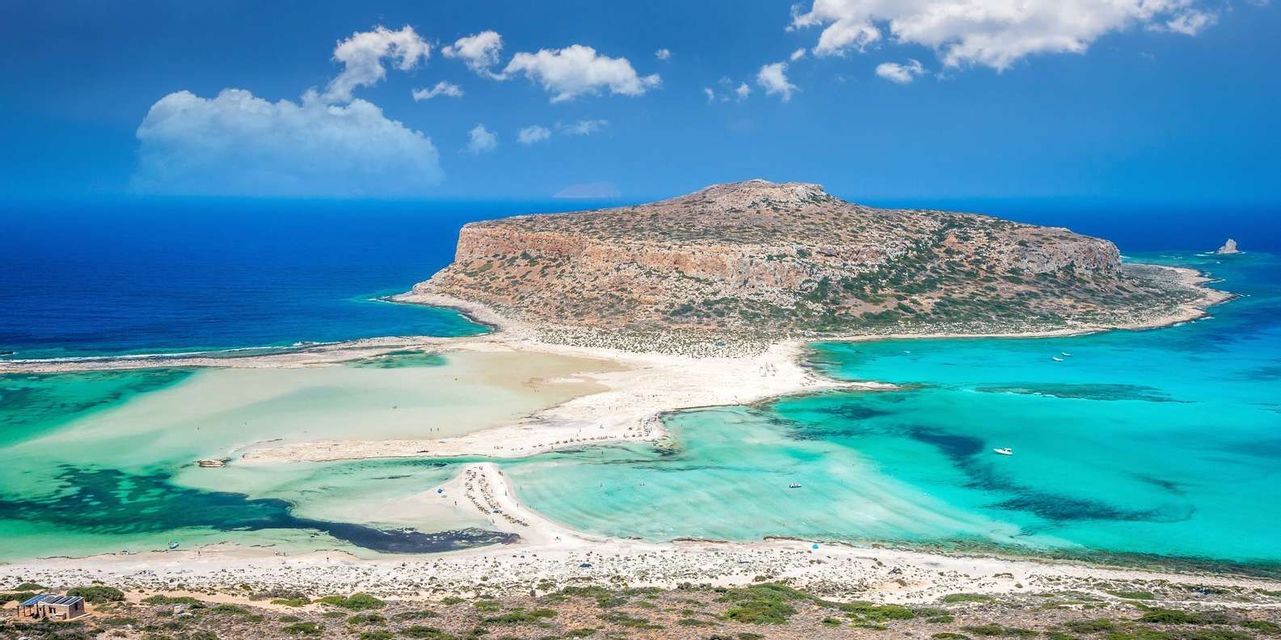Vista aerea di un'isola rocciosa con una lingua di sabbia che separa una laguna turchese dal mare blu intenso.