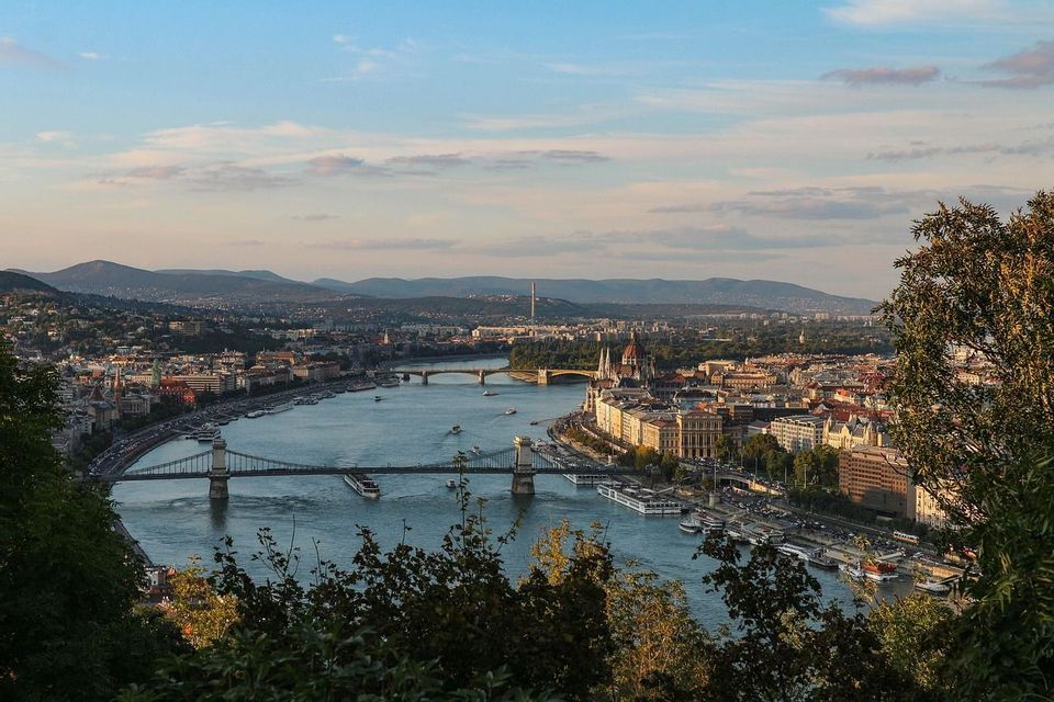 Una vista panoramica di una città costruita lungo un ampio fiume, con diversi ponti, barche e dolci colline in lontananza sotto un cielo tenue.