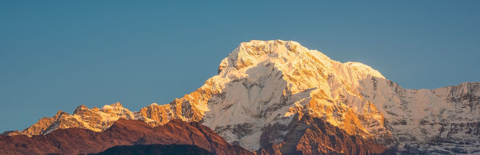 Una grande cima montuosa coperta di neve illuminata dalla luce dorata del sole contro un cielo azzurro e limpido.